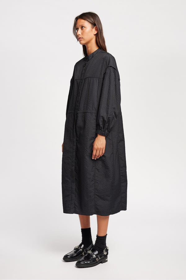 Front angle of Model wearing the Parapet Dress in Jet Black. A lightweight seersucker nylon, showing band collar and adjustable front waist tie.