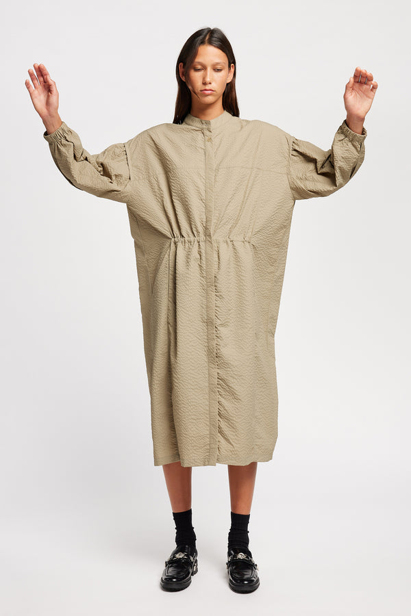 Front of Model wearing the Parapet Dress in Stone/beige. A lightweight seersucker nylon, showing band collar and adjustable front waist tie.