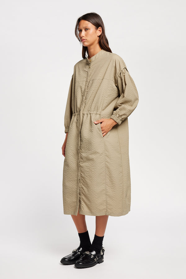 Front of Model wearing the Parapet Dress in Stone/beige. A lightweight seersucker nylon, showing band collar and adjustable front waist tie.