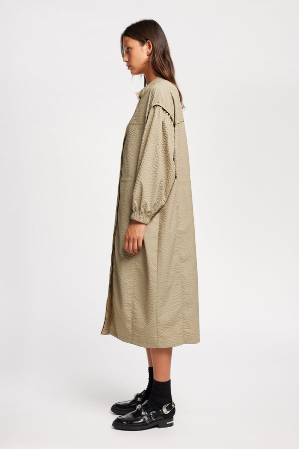 Side of Model wearing the Parapet Dress in Stone/beige. A lightweight seersucker nylon, showing band collar and adjustable front waist tie.