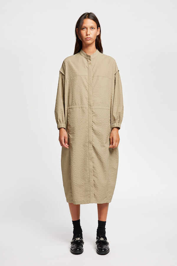 Front of Model wearing the Parapet Dress in Stone/beige. A lightweight seersucker nylon, showing band collar and adjustable front waist tie.