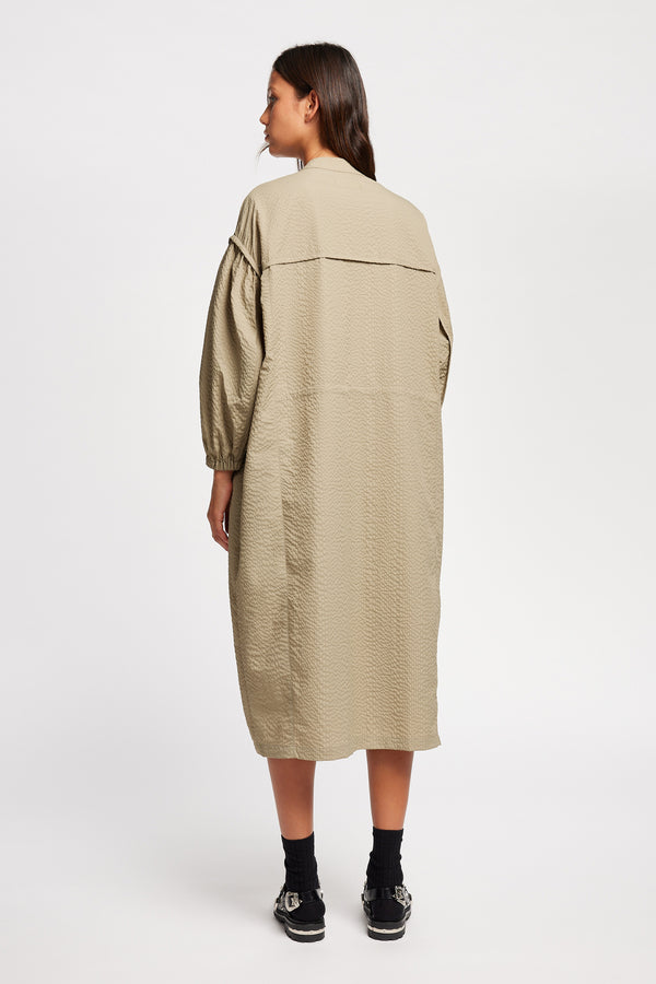 Back of Model wearing the Parapet Dress in Stone/beige. A lightweight seersucker nylon, showing band collar and adjustable front waist tie.