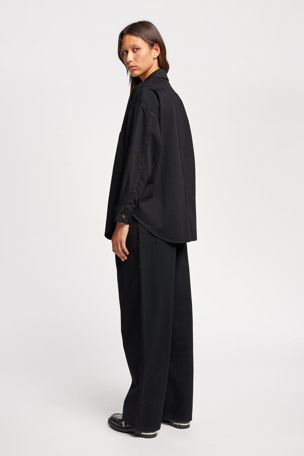 Back angle view of model wearing the Region oversized denim shirt in Black, featuring utility chest pockets.