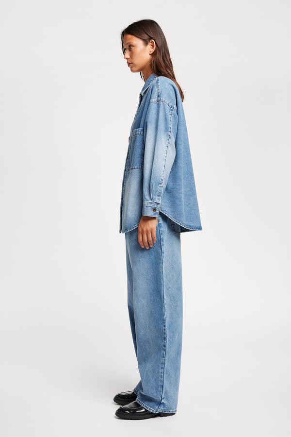 Side view of model wearing the Region oversized denim shirt in mid-blue, featuring utility chest pockets.