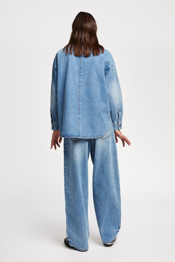 Back view of model wearing the Region oversized denim shirt in mid-blue, featuring utility chest pockets.