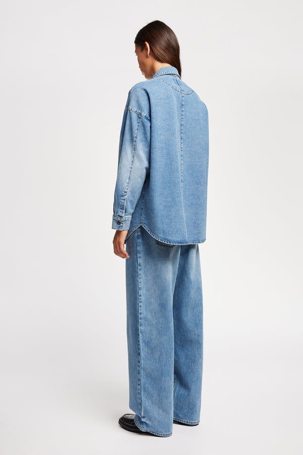 Back angle view of model wearing the Region oversized denim shirt in mid-blue, featuring utility chest pockets.