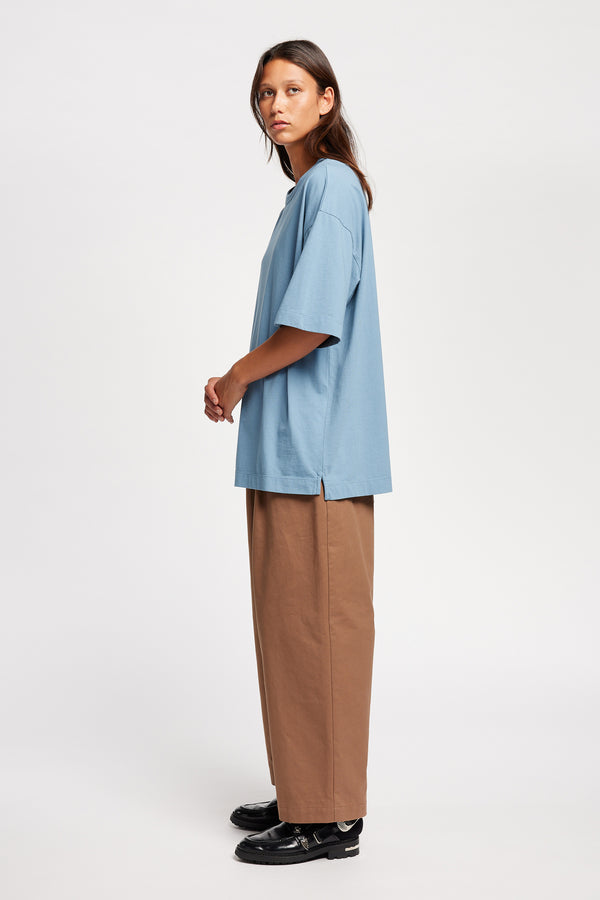 Side of Model wearing Seleno pants in clay brown with a relaxed, tapered leg and pleat detailing, styled with Monolith Tee in Blue.