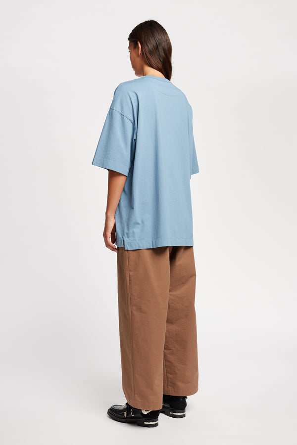 Back of Model wearing Seleno pants in clay brown with a relaxed, tapered leg and pleat detailing, styled with Monolith Tee in Blue.
