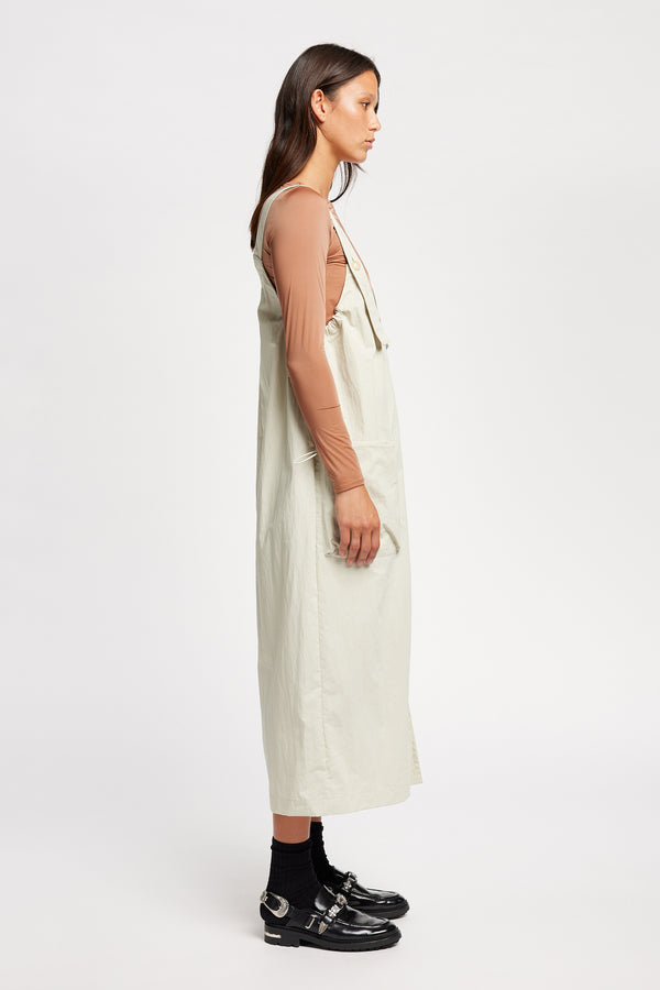 Side of the model wearing the Shoreline full-length pinafore dress in bone white with deep round neckline and front patch pockets, a relaxed-fit silhouette. 
