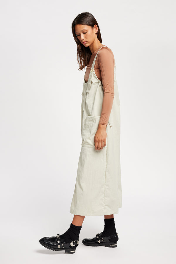 Side of the model wearing the Shoreline full-length pinafore dress in bone white with deep round neckline and front patch pockets, a relaxed-fit silhouette. 