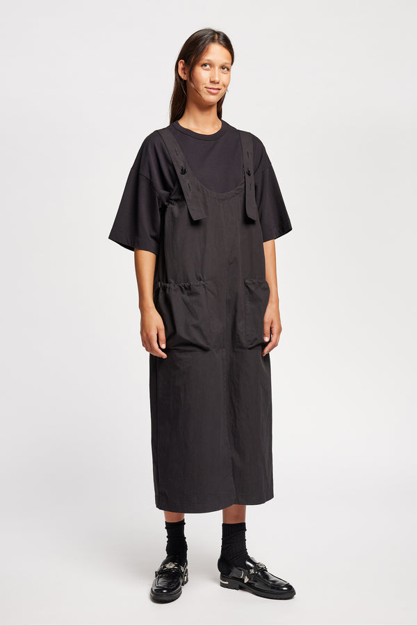 Font of the model wearing the Shoreline full-length pinafore dress in black with deep round neckline and front patch pockets, a relaxed-fit silhouette. 