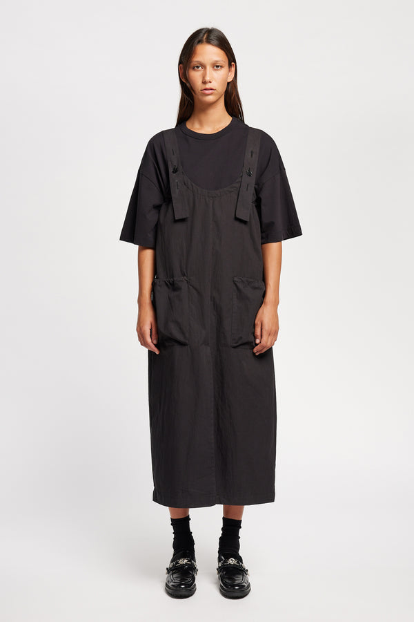 Font of the model wearing the Shoreline full-length pinafore dress in black with deep round neckline and front patch pockets, a relaxed-fit silhouette. 