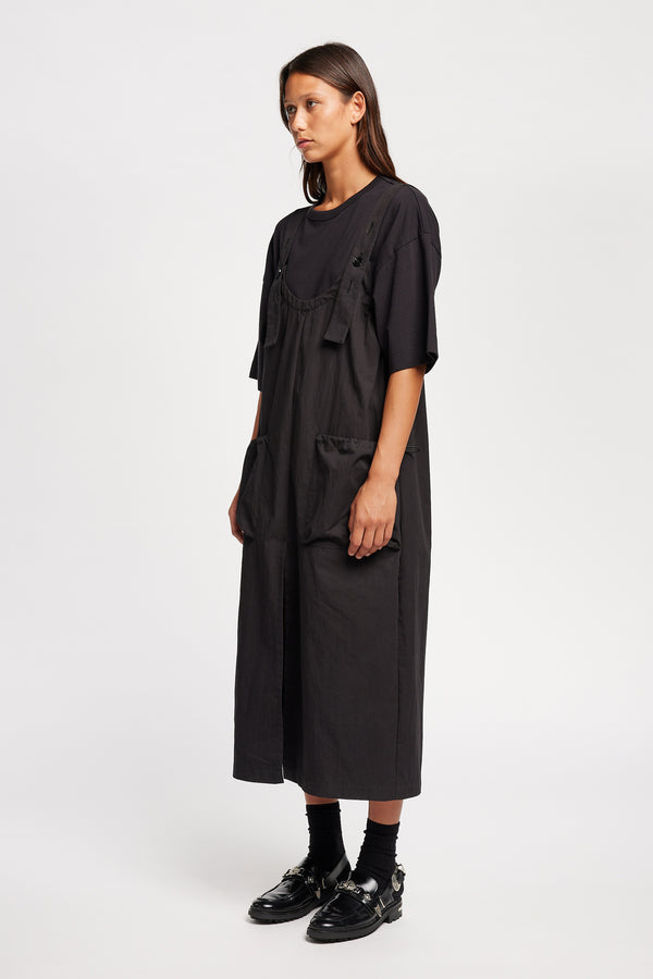 Font of the model wearing the Shoreline full-length pinafore dress in black with deep round neckline and front patch pockets, a relaxed-fit silhouette. 