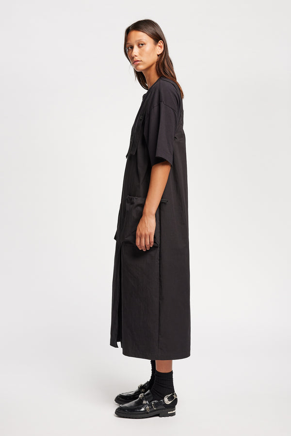 Side of the model wearing the Shoreline full-length pinafore dress in black with deep round neckline and front patch pockets, a relaxed-fit silhouette. 