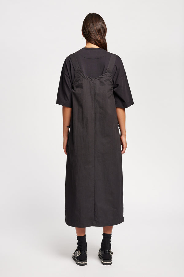 Back of the model wearing the Shoreline full-length pinafore dress in black with deep round neckline and front patch pockets, a relaxed-fit silhouette. 