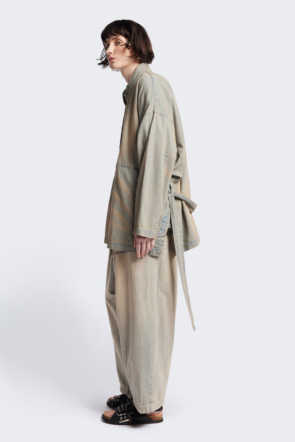 Side of the unisex Subterra Wide wrap shawl collar jacket with kimono-style sleeves, crafted from cotton denim, featuring front patch pockets and a waist tie to cinch the loose silhouette. Available in 5 sizes.  