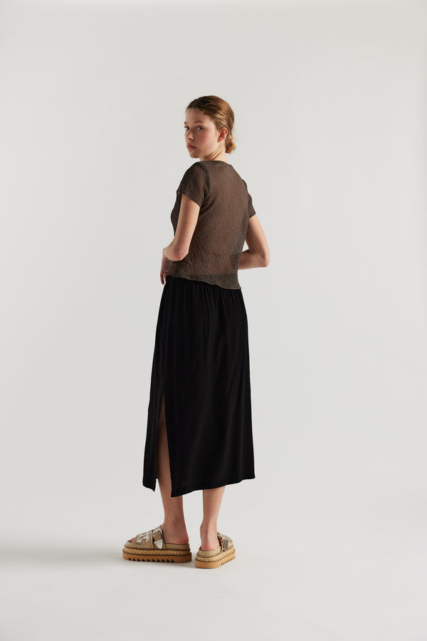 Back view on model of Mid-length slip skirt in black soft viscose-rayon. Ft. elasticated gathered waist with self-tie pull and side split visible.