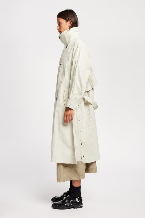 Side view of model wearing the Volt Trench in Bone collar up. A lightweight double-breasted trench coat with flap pockets
