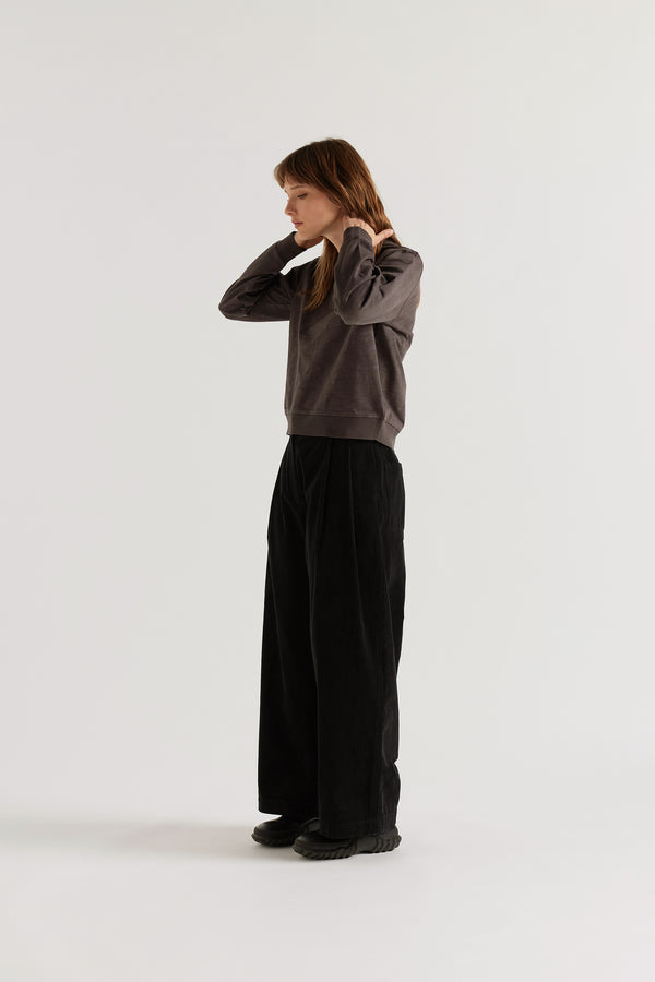 Three-quarter view of the black Willow Cord Pant showing wide balloon-shaped leg, pleat-front detail and relaxed semi-tailored corduroy silhouette