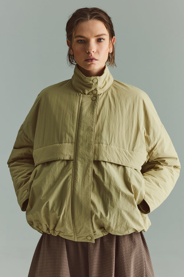 Front of female model in the Sand Beige Surge Quilted Jacket worn open, showing relaxed silhouette, military tab collar and storm flap pocket detailing