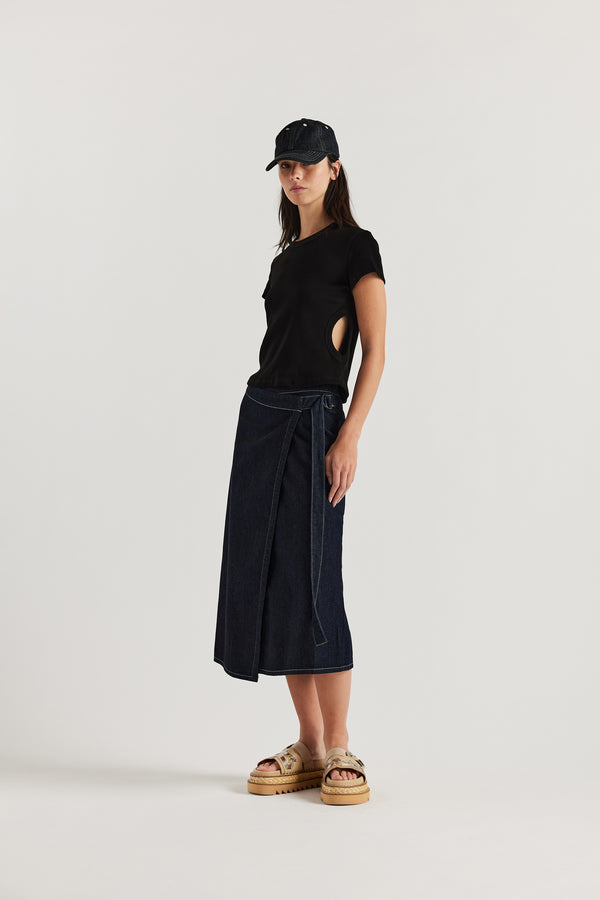 Female model wears KLOKE vital wrap skirt in indigo denim with contrast white stitch, styled with matching denim cap. black side-cut tee and beige slide. Three quarter view