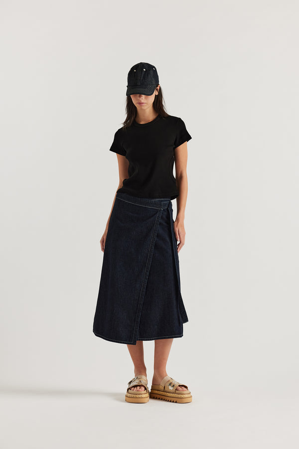 Female model wears KLOKE vital wrap skirt in indigo denim with contrast white stitch, styled with matching denim cap. black side-cut tee and beige slide. Front view